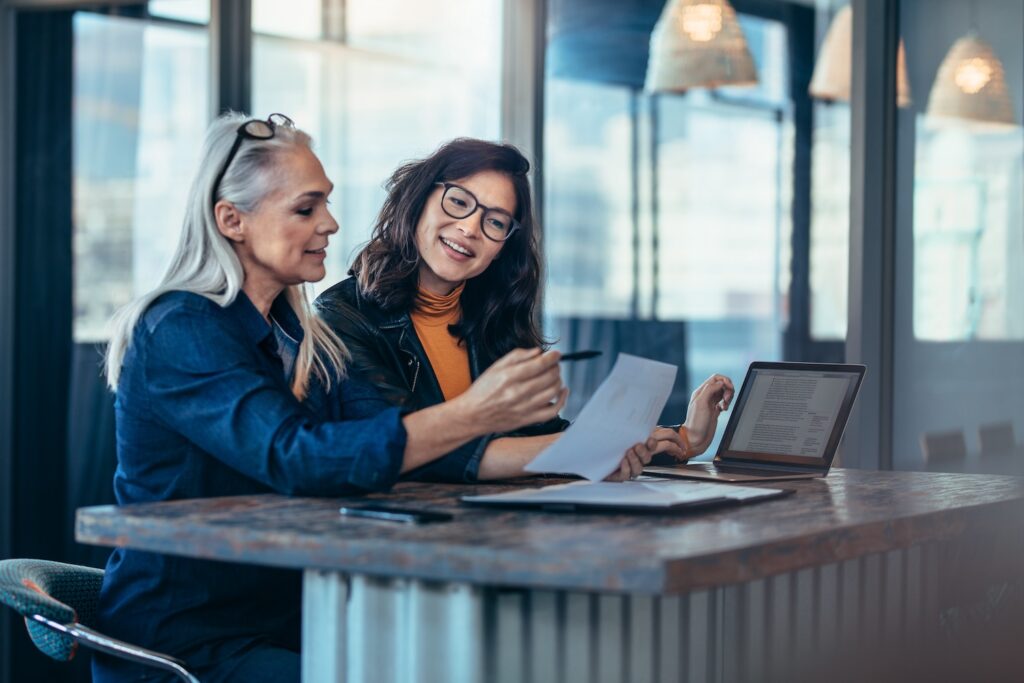 Two women reviewing papers while sitting in front of a laptop at a high counter top