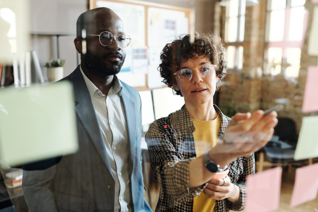 Young businesswoman discussing plans on sticky notes hanging on glass wall together with her colleague at office