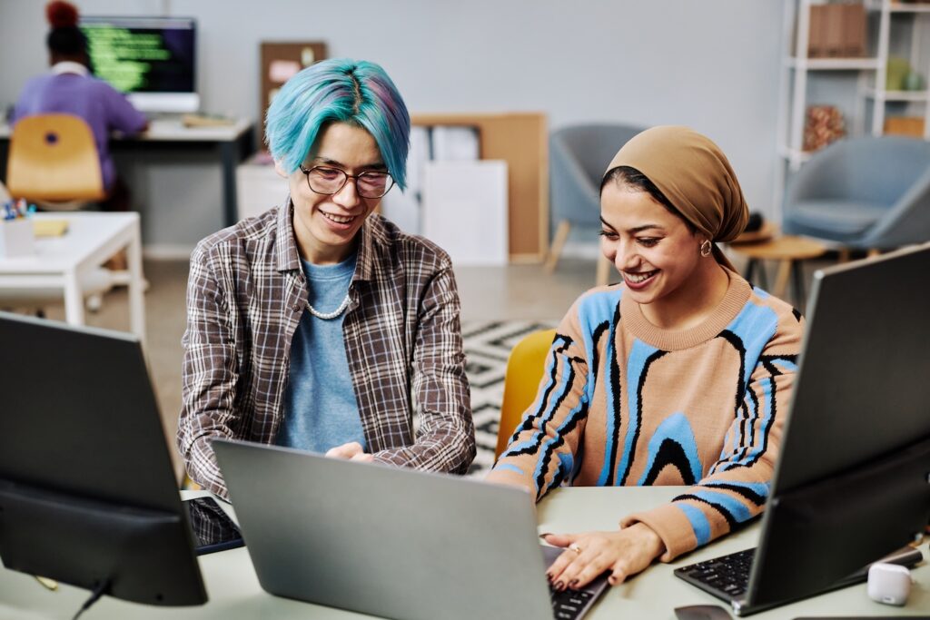 Portrait of two young software developers using computers in office and smiling happily enjoying work