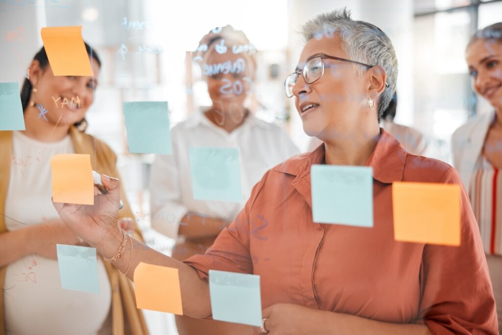 Woman with gray hair surrounded by colleagues as they review and write on sticky notes on a glass wall