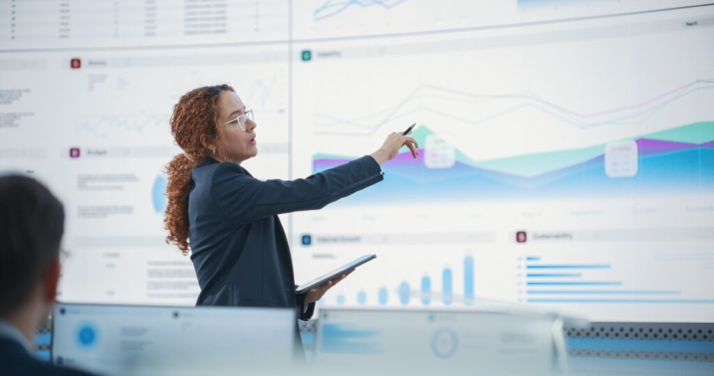 Woman gives presentation in front of large screen showing data visualized in blue, green and purple charts