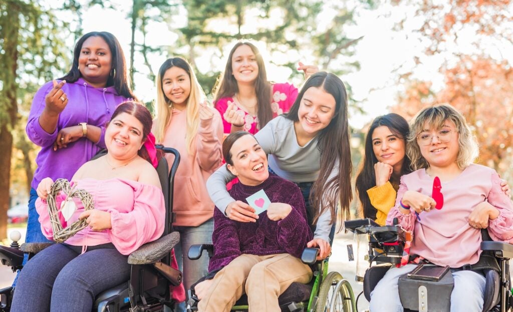 Group of multiracial women, some using wheelchairs smiling and making heart symbols