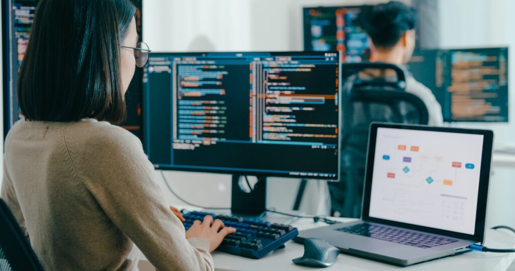 Asian woman coding at a large monitor and laptop