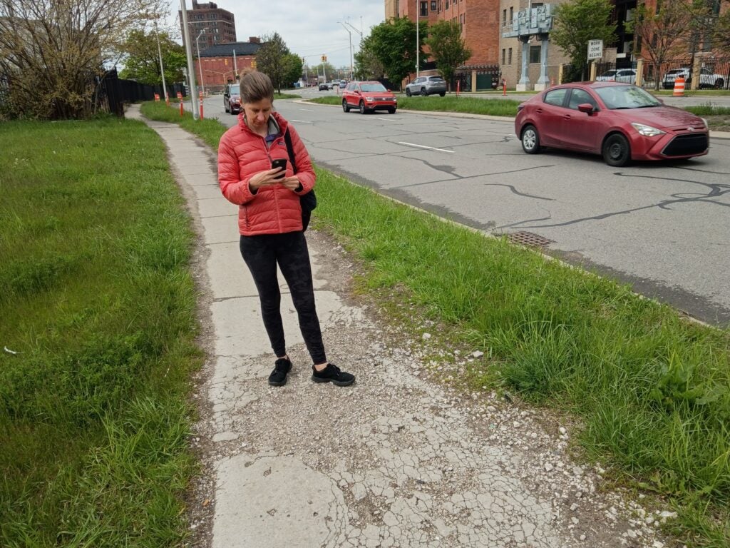 Bridget Scallen stands on a crumbling stretch of Martin Luther King Jr Blvd between Woodward & Cass with her phone as she documents cracked and weathered concrete on May 1, 2026. Photo credit: Bridget Scallen.