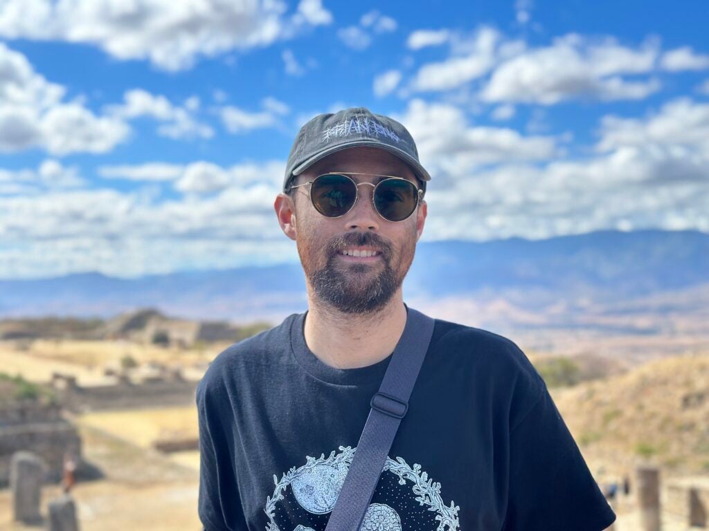 Sean Isamu Johnson smiles in front of a vast, mountainous vista and ancient ruins.